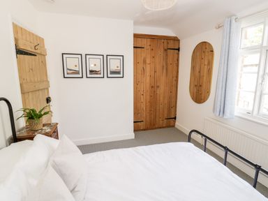 A bedroom with a bed a wooden door a wooden closet a bedside table with a plant three framed artworks a wall mirror and a window with curtains at Ross Cottage in Broadway