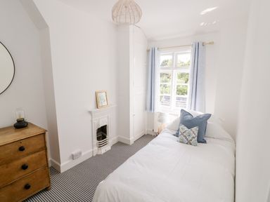 A bedroom with a single bed white bedding and blue pillows a wooden chest of drawers and a window with blue curtains at Ross Cottage Broadway
