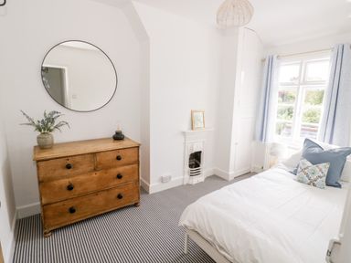 A bedroom with a wooden chest of drawers a round mirror a bed with pillows and a window with curtains at Ross Cottage in Broadway
