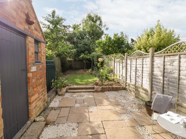 A patio with stone tiles and white gravel next to a brick building and a garden with green grass and trees at Ross Cottage in Broadway