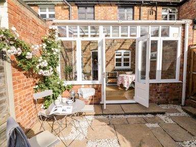 A patio with a small table and chairs outside a brick house with a glass conservatory door open at Ross Cottage in Broadway