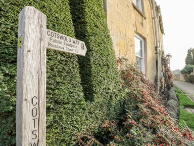A wooden signpost for Cotswold Way public footpath next to a stone building with windows and a hedge with red berries at Ross Cottage in Broadway