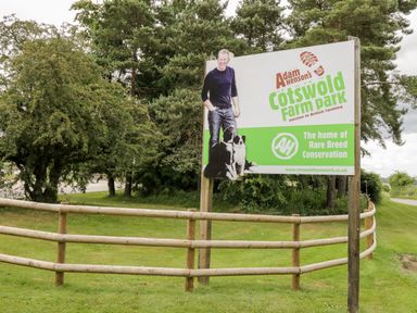 A sign for Adam Henson's Cotswold Farm Park next to a wooden fence and trees in the background