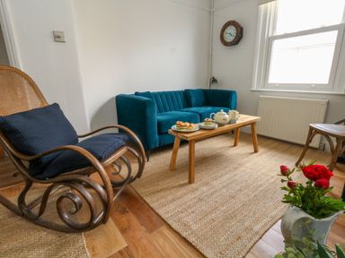 A living room with a blue sofa a wooden rocking chair a wooden coffee table with tea and pastries a clock on the wall and a plant at 26 High West in Weymouth