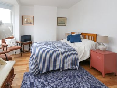 A bedroom with a wooden bed with blue and white bedding a blue patterned rug two armchairs a small television on a wooden table and a pink bedside cabinet at 26 High West in Weymouth