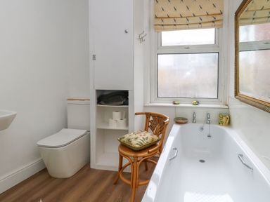 A bathroom with a bathtub a toilet a wooden chair with a cushion a built-in shelf and a window with a blind at 26 High West in Weymouth