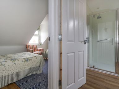 A bedroom with a bed and striped armchair next to a window and an adjacent bathroom with a glass shower enclosure at 26 High West in Weymouth