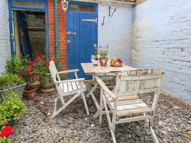 An outdoor patio area with a wooden table and two chairs on a gravel surface with potted plants and blue doors at 26 High West in Weymouth