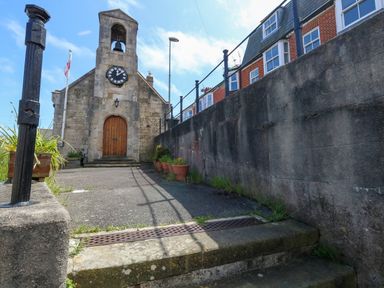 An exterior view of a stone building with a clock and bell tower beside a concrete path and brick houses at 26 High West in Weymouth