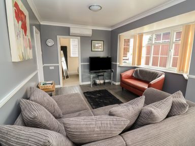 A living room with a large sofa and cushions a red armchair a television on a glass stand and windows at Swiss Cottage in Wilsthorpe near Bridlington