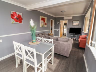A dining area with a table and chairs next to a living room with sofas a television and wall art at Swiss Cottage Wilsthorpe near Bridlington