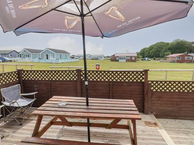 A wooden picnic table with an umbrella and a folding chair on a deck overlooking a grassy area with houses at Swiss Cottage in Wilsthorpe near Bridlington