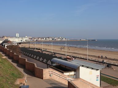 A beach with people walking near rows of small beach huts and a promenade at Swiss Cottage in Wilsthorpe near Bridlington