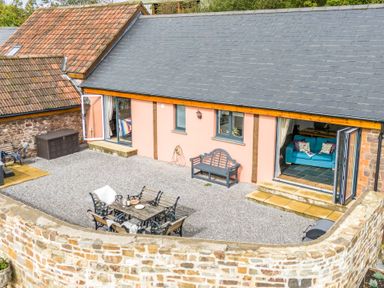 An outdoor seating area with a table and chairs and a bench outside a building with open doors at The Farm Office in Bampton Devon