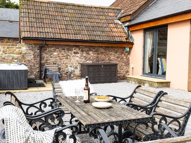 An outdoor patio area with a wooden table and chairs set on gravel in front of a stone wall and a building with sliding glass doors at The Farm Office in Bampton Devon