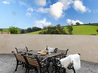 An outdoor patio with metal and wood table and chairs on gravel with green hills and trees in the background at The Farm Office in Bampton Devon