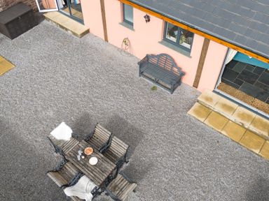 An outdoor patio with a wooden table and chairs set on gravel next to a peach colored building with windows and a bench at The Farm Office in Bampton Devon