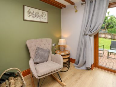 A corner of a room with a beige rocking chair a side table with a lamp and a basket of firewood next to a glass door with grey curtains at Pondarosa in Newbridge-On-Wye