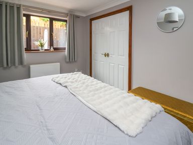 A bedroom with a bed covered in a white textured blanket and throw blanket a window with curtains a double door closet and a round mirror on the wall at Pondarosa in Newbridge-On-Wye