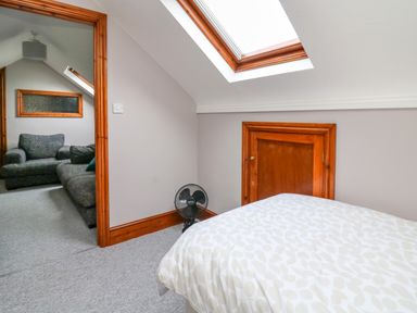 A bedroom with a bed under a skylight window and a small door on the wall next to a fan and an adjacent room with two grey sofas at Pondarosa in Newbridge-On-Wye