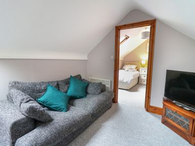 A sitting area with a gray sofa and teal cushions a television on a wooden stand and a view into a bedroom at Pondarosa in Newbridge-On-Wye