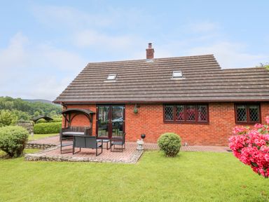 A brick house with a tiled roof and skylights a patio with outdoor seating and a garden with bushes and flowers at Pondarosa in Newbridge-On-Wye