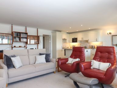 A living room with a beige sofa and two red armchairs facing a kitchen with white cabinets at 14 Burgh Island Causeway in Bigbury-On-Sea