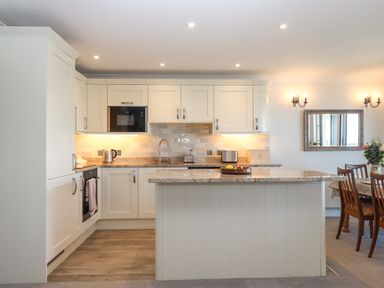 A kitchen with cabinets appliances and an island with a fruit bowl next to a dining area with wooden chairs and a table at 14 Burgh Island Causeway in Bigbury-On-Sea