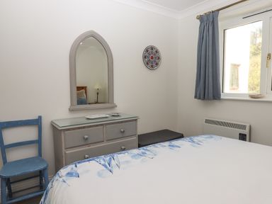 A bedroom with a bed a blue chair a dresser with a mirror and a window with blue curtains at 14 Burgh Island Causeway in Bigbury-On-Sea