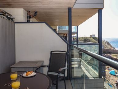 A balcony with a round table set with croissant and juice glasses and a chair overlooking a beach at 14 Burgh Island Causeway in Bigbury-On-Sea