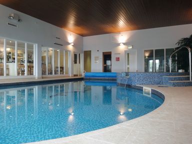 An indoor swimming pool with steps and a small waterfall next to seating area with glass walls showing dining tables and chairs at 14 Burgh Island Causeway in Bigbury-On-Sea