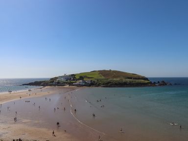 A sandy beach with people walking and swimming with an island and buildings in the background at 14 Burgh Island Causeway in Bigbury-On-Sea
