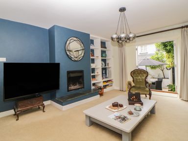 A living room with a TV on a stand a fireplace a round mirror built-in shelves an armchair a coffee table and an open door to a garden at Lower Culvermead in Chaddlehanger near Tavistock