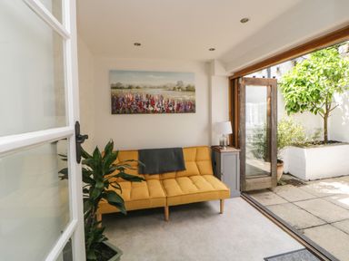 A small sitting room with a mustard yellow sofa a gray throw a small cabinet with a lamp a plant and a door open to a patio at Lower Culvermead in Chaddlehanger near Tavistock