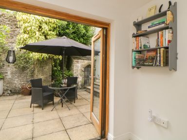 A patio with a table umbrella and two chairs seen through open doors and a wall-mounted bookshelf inside Lower Culvermead in Chaddlehanger near Tavistock