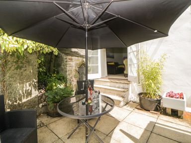 A patio with a round glass table under a large umbrella black chairs plants and open doors leading inside at Lower Culvermead in Chaddlehanger near Tavistock