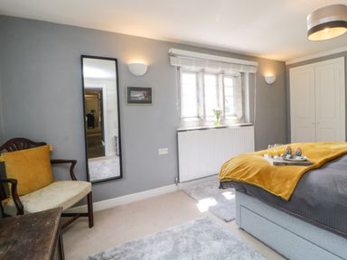 A bedroom with a bed covered in gray and yellow bedding a wooden chair with a yellow cushion a tall mirror and a window at Lower Culvermead in Chaddlehanger near Tavistock