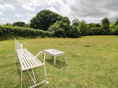 A white metal bench and a wooden table on a grassy field with trees and bushes in the background at Lower Culvermead in Chaddlehanger near Tavistock