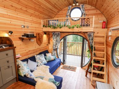 A small wooden living room with a blue sofa cushions a loft area and circular glass doors opening to a balcony at The Kites Nest in Pont-rhyd-y-groes near Pontrhydfendigaid Snowdonia
