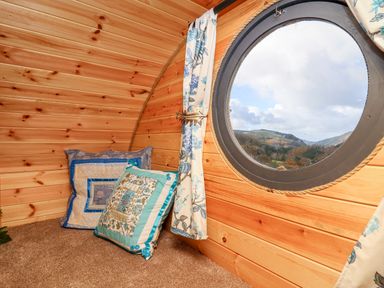 A wooden interior corner with two patterned cushions on a carpeted floor and a round window with floral curtains at The Kites Nest in Pont-rhyd-y-groes near Pontrhydfendigaid Snowdonia