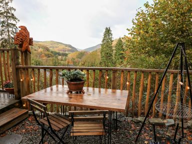 An outdoor wooden table with a plant and two chairs on a deck with a wooden railing and a carved eagle sculpture surrounded by trees at The Kites Nest in Pont-rhyd-y-groes near Pontrhydfendigaid Snowdonia
