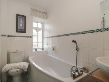 A bathroom with a bathtub shower, toilet with a white mat, framed artwork, and a window at Teal House in Lyme Regis