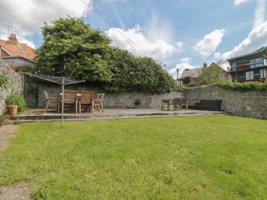 A backyard with a green lawn a rotary clothesline wooden and metal outdoor tables and chairs against a stone wall at Teal House in Lyme Regis