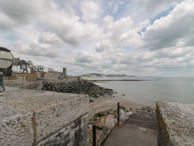 A stone walkway and steps leading to a rocky shoreline with buildings and hills in the background at Teal House in Lyme Regis