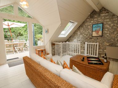 A sitting area with a wicker sofa and table near a stone wall and glass door opening to an outdoor dining space at Tan y Craig in Solva