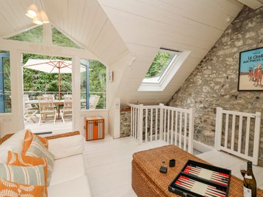 A sitting area with a white sofa and striped orange cushions a wicker coffee table with a backgammon game and a bottle and glass of wine a slanted ceiling with a skylight and a stone wall at Tan y Craig in Solva