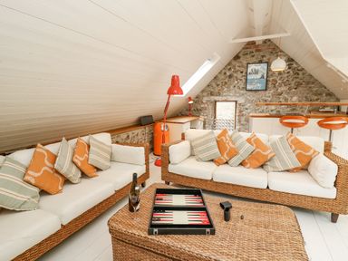 A living area with two wicker sofas with cushions a wicker coffee table with a backgammon game wine glass and bottle a stone wall with a framed poster and kitchen bar with stools at Tan y Craig in Solva