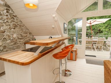A kitchen area with stone wall wooden countertops two orange bar stools and a glass door opening to an outdoor dining area with wooden chairs and a table under an umbrella at Tan y Craig in Solva