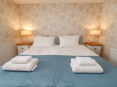 A bedroom with a double bed two white pillows blue blanket folded white towels bedside tables with lamps wallpaper with seaweed pattern at Tan y Craig in Solva
