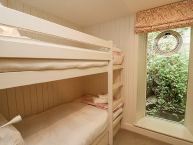 A bedroom with white bunk beds and a window with a circular opening showing greenery outside at Tan y Craig in Solva